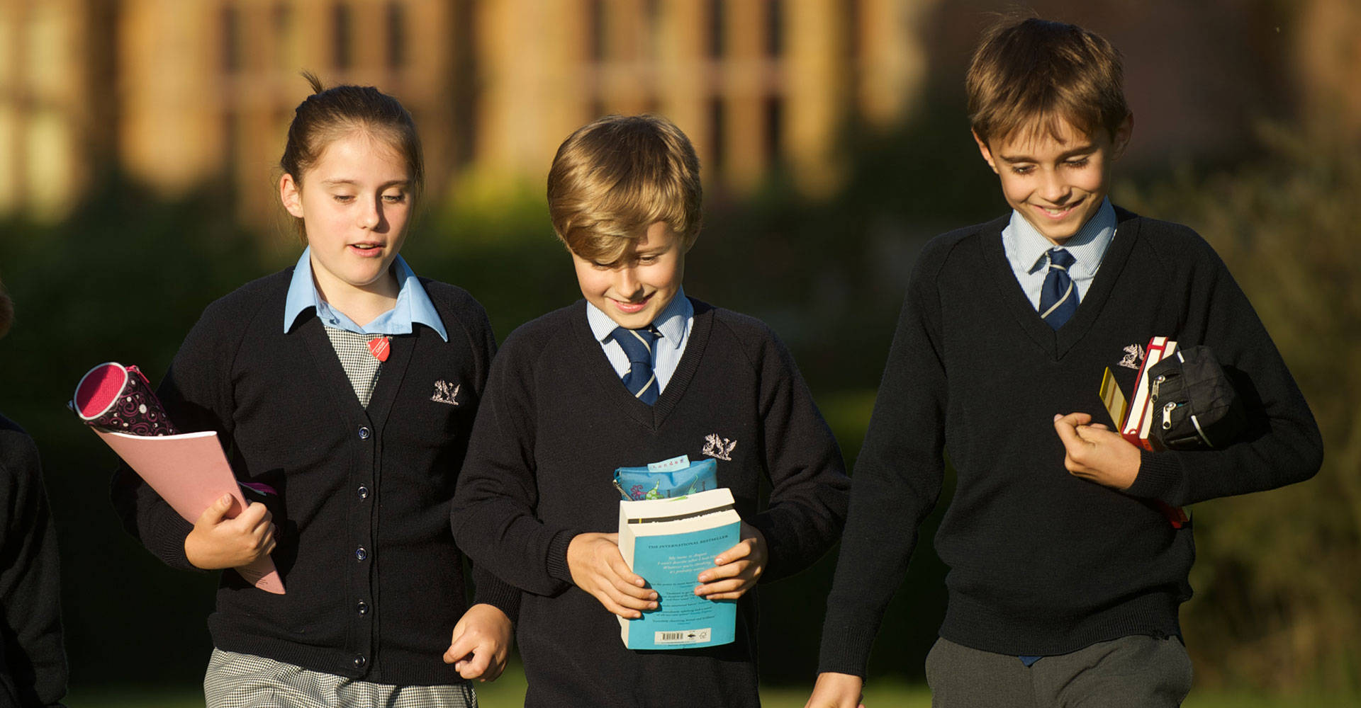 Three children in school uniforms walk outside, holding books and smiling. The uniforms include dark sweaters, ties, and shirts. They appear to be enjoying a conversation in a green area with blurred trees in the background.