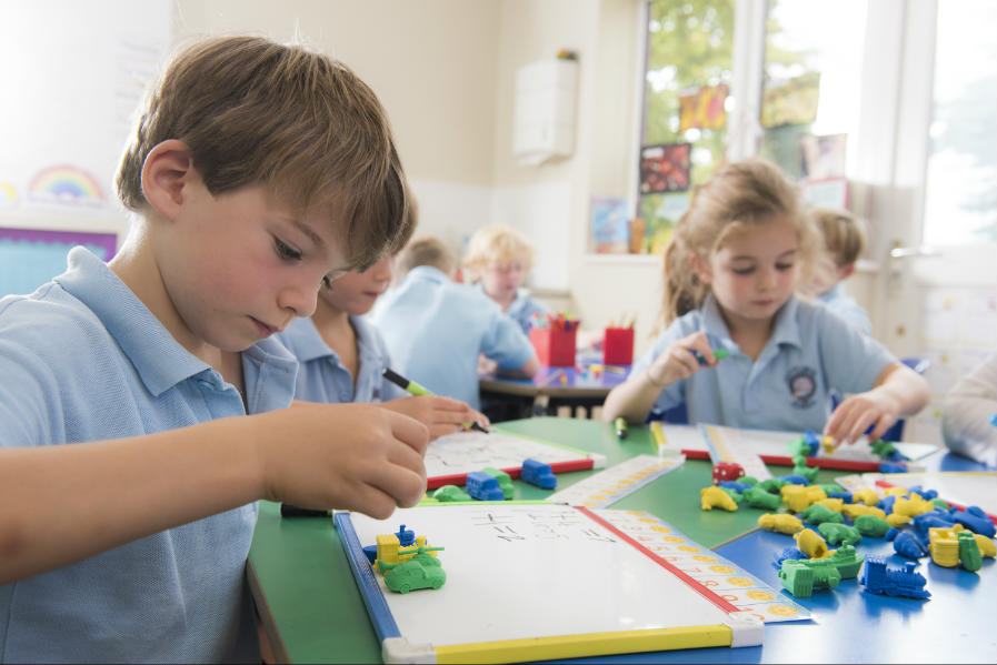 Children in a classroom engage in activities using colored geometric blocks on their desks. They are wearing blue uniforms and focused on building shapes and patterns. Bright windows and educational materials fill the background.