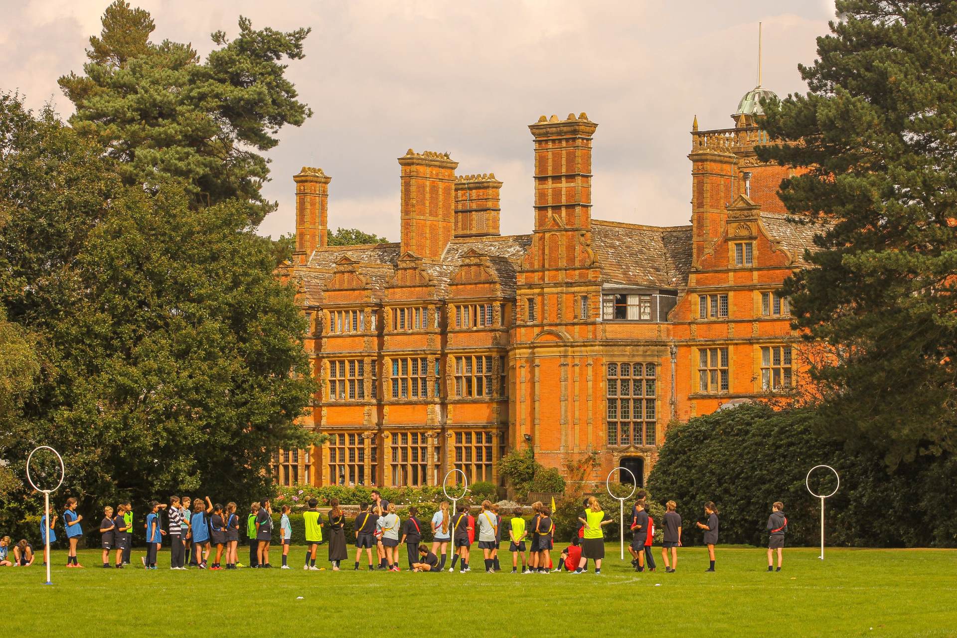 A group of people gather on a grassy field with Quidditch hoops in front of a large, historic red-brick building surrounded by trees. Some wear team jerseys, indicating they are preparing for a Quidditch match. The setting is sunny and vibrant.