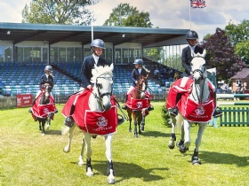 Four riders on white horses, all wearing helmets and black attire, parade on a grassy field. The horses are draped in red blankets with a white logo. An empty grandstand and a British flag are visible in the background under a partly cloudy sky.