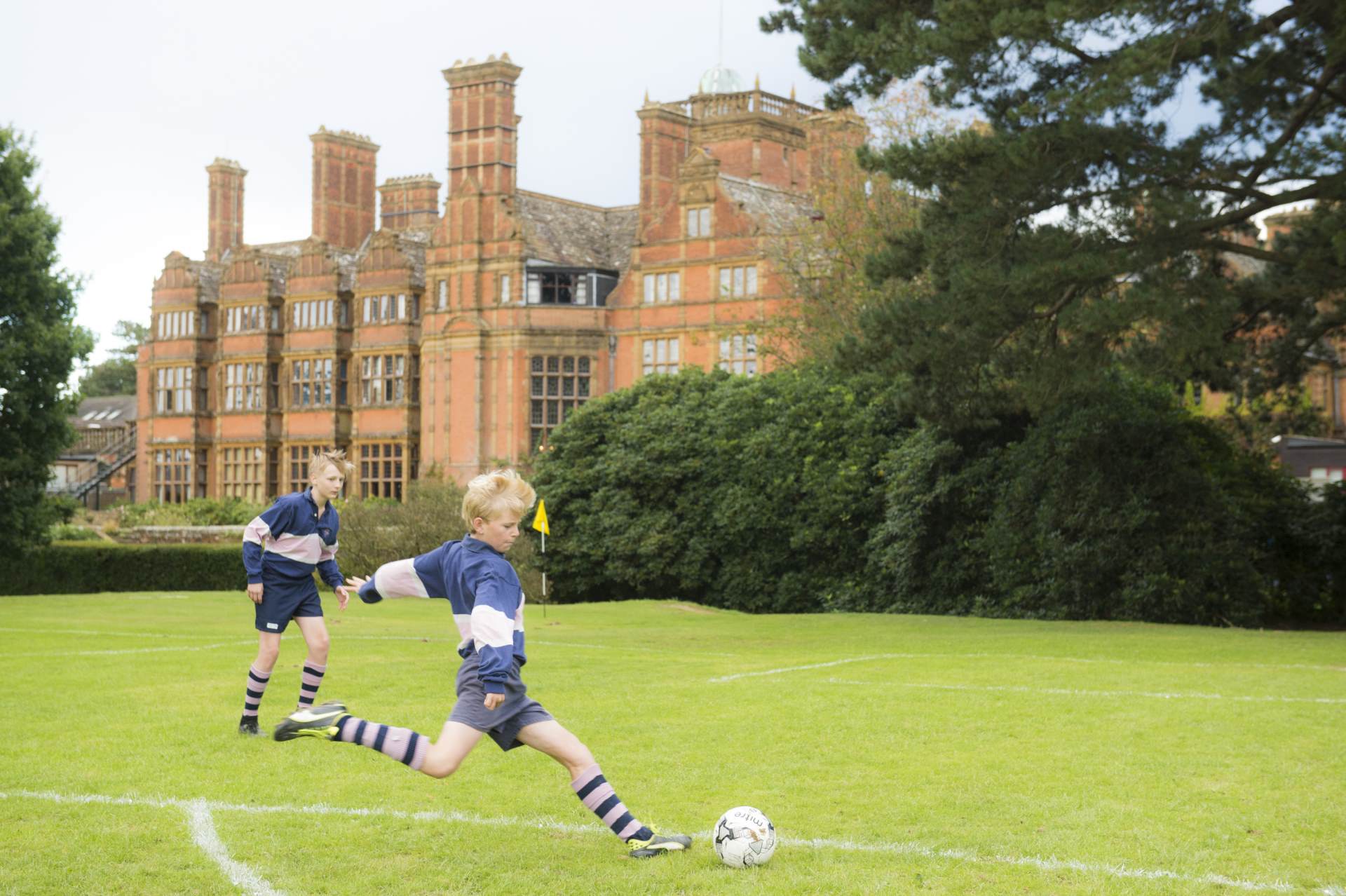 cottesmore school male pupils football kit playing on pitch field about to shoot the ball with school building in background