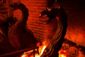 Two wrought iron dragon-shaped andirons sit in front of a glowing fireplace. The scene is warmly lit by the fire, highlighting the intricate details of the dragon heads against a backdrop of red brick.