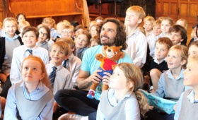 A man seated in the middle of a group of young children, holding a teddy bear. The children are wearing school uniforms with ties or checkered dresses, smiling and looking in various directions. Wooden furniture is visible in the background.