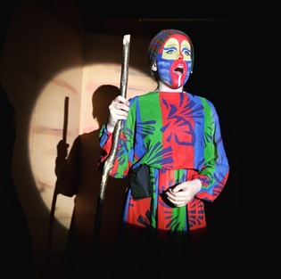 cottesmore school female pupil in colourful costume with face paint singing on stage under spotlight holding wooden staff