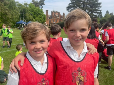 Two children wearing red sports bibs stand arm-in-arm, smiling. They are outdoors on a grassy field with other children in the background, and a large building surrounded by trees is visible.