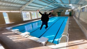 A person in a black outfit is jumping into an empty indoor swimming pool with arms raised. The pool area is well-lit with sunlight streaming through large windows. The ceiling is curved with blue panels.