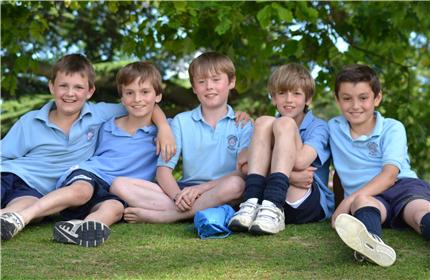 Five boys in light blue shirts and shorts sit closely on grass, smiling under a leafy tree. They appear relaxed and are enjoying the outdoors together, with arms around each other.