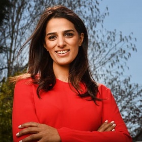 A woman with long dark hair wearing a red top stands outdoors with trees and a blue sky in the background, smiling confidently with arms crossed.