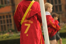 A person wearing a red coat with POTTER 7 and a star printed on the back stands next to a white pole. Two other individuals, slightly blurred, are in the background. They appear to be outdoors in a garden setting.