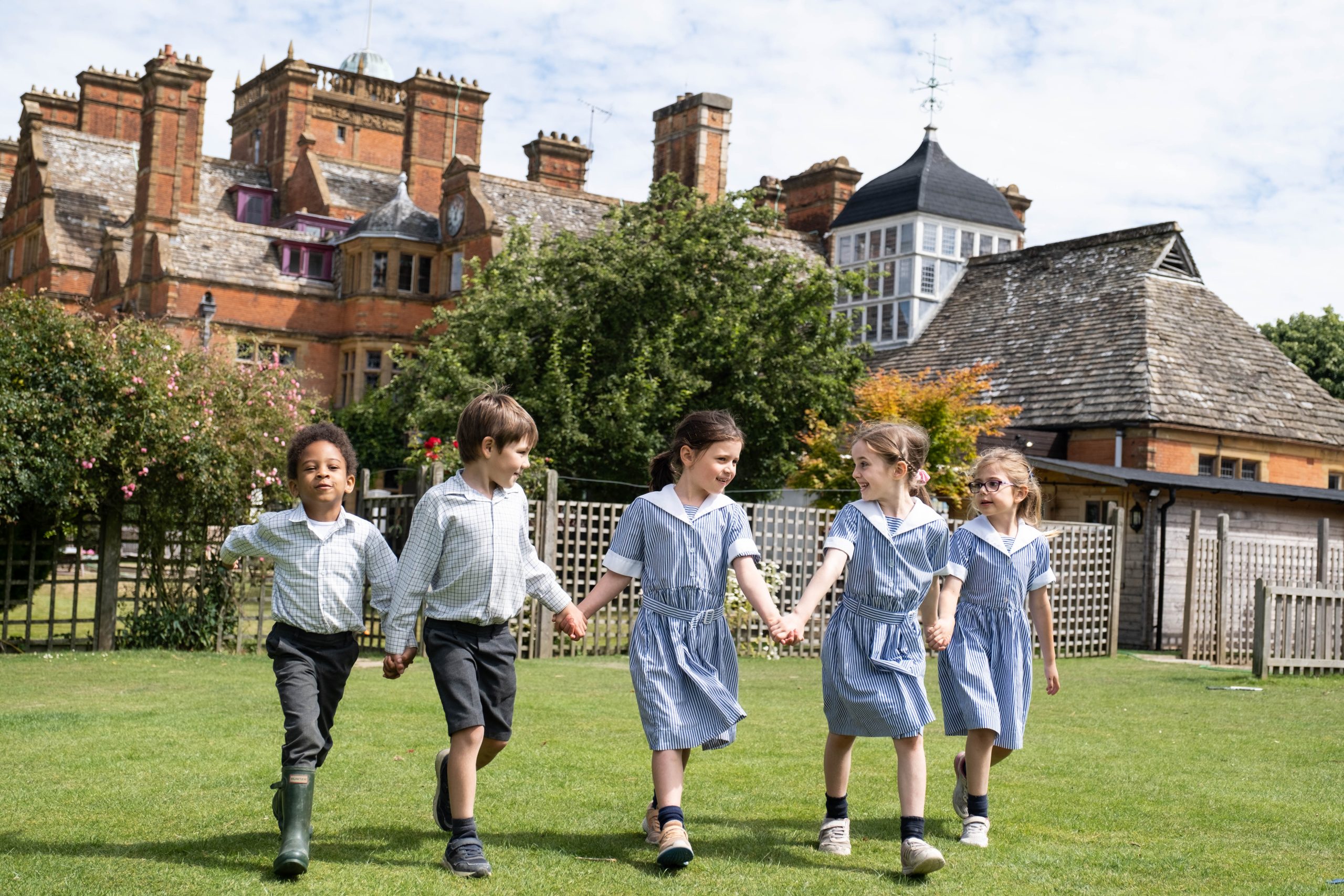 Five children in school uniforms walk hand in hand on a grassy field. They appear happy and are walking in front of a large, historic building with brick walls and chimneys. The sky is partly cloudy.