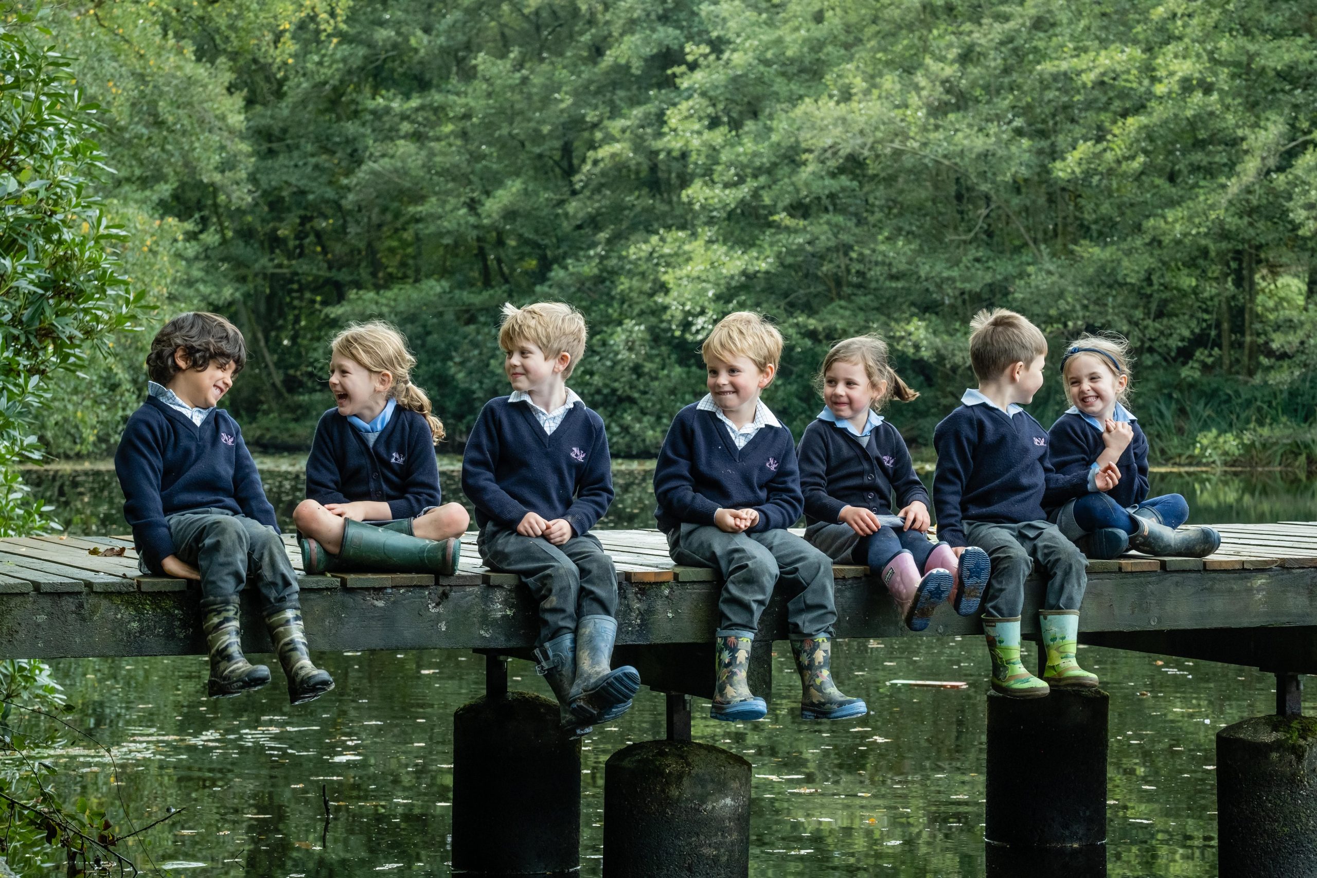 Seven young children in school uniforms sit side by side on a wooden dock over a lake, surrounded by lush green trees. They wear rain boots and are smiling and chatting with each other, creating a joyful and playful scene.